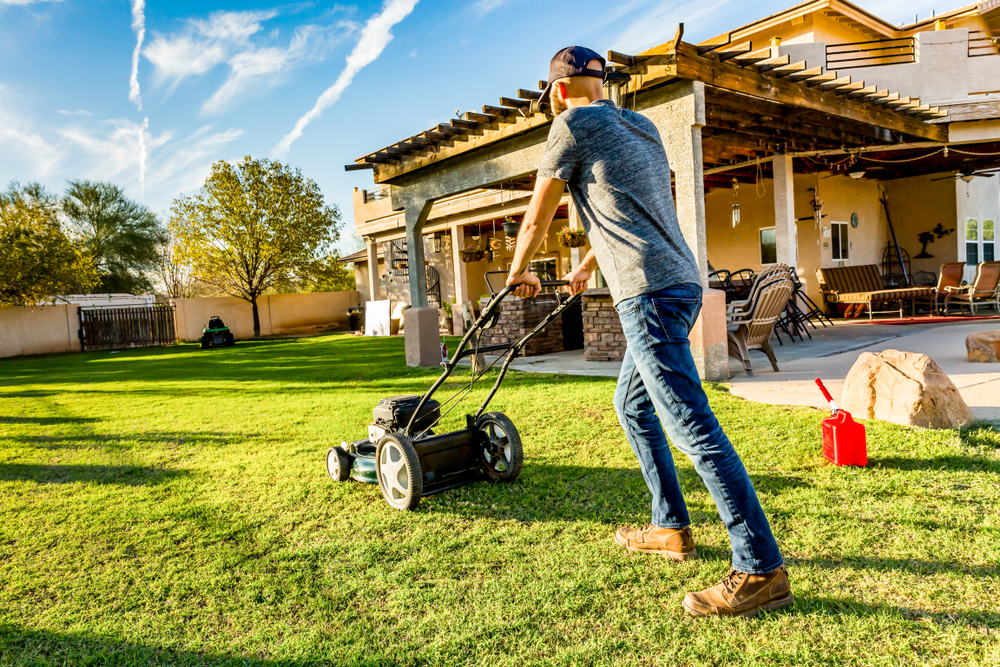 Man in baseball cap mowing green lawn on bright summer day in backyard. Red gasoline can sits nearby on grass ready to refuel mower.