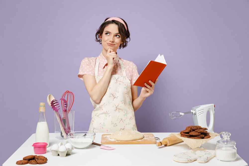 woman in pink apron work at table kitchenware holding recipe cook book look aside isolated on pastel violet background Process cooking food concept